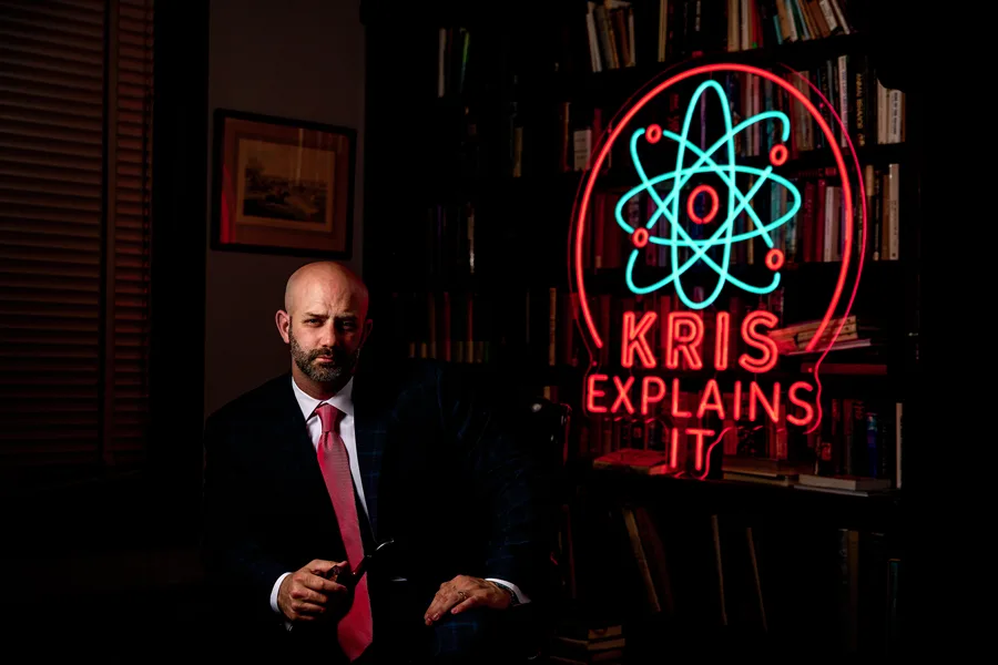 Kris's co-host seated beside the neon Kris Explains It sign